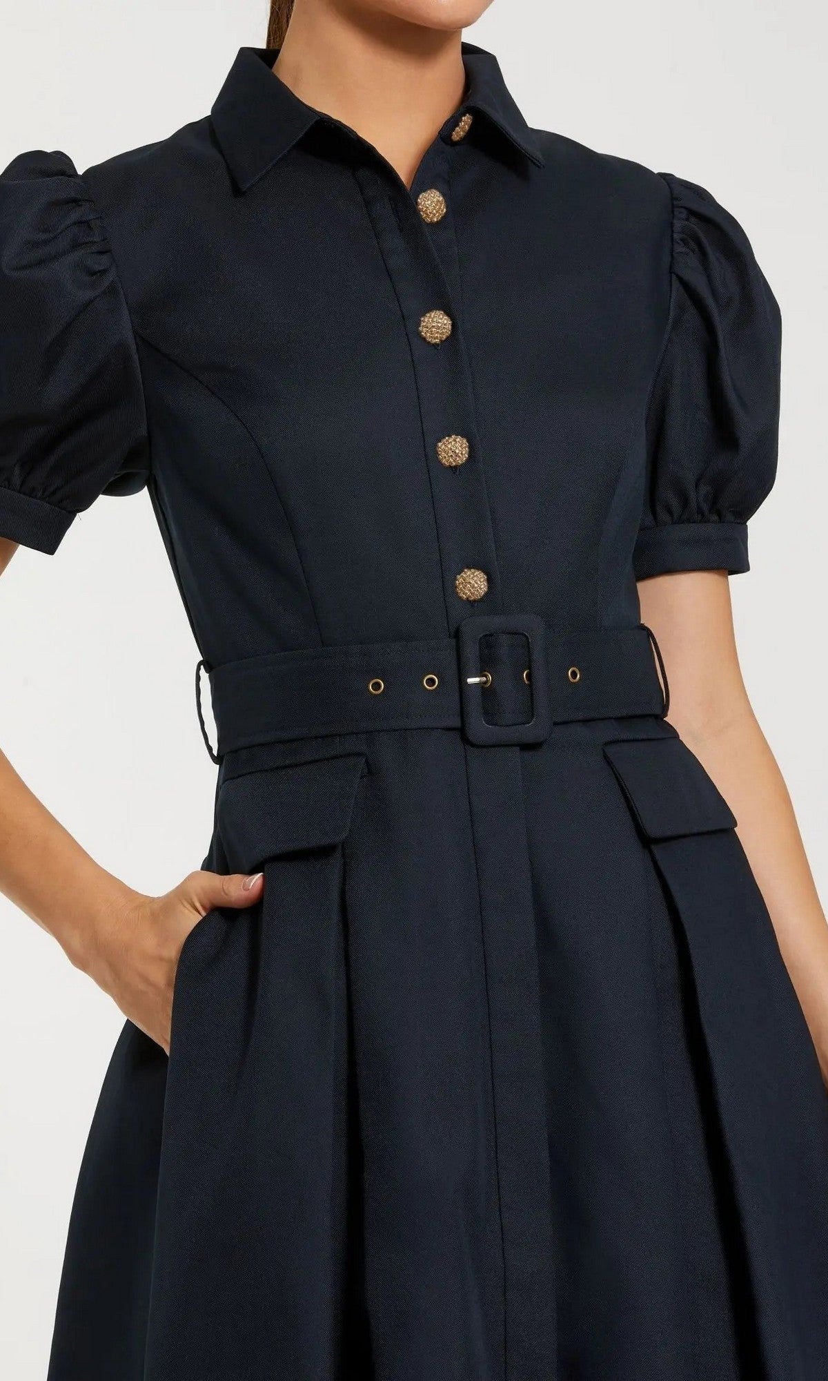 A person wearing the Mac Duggal 69097 Knee Length Belted Shirt Dressโfeaturing a dark hue, short puff sleeves, gold buttons, a collared neckline, and flap pocketsโposes with one hand in a pocket against a plain background.